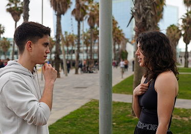 Joven con sudadera blanca y mujer con ropa deportiva negra conversan en un parque con palmeras y un edificio de cristal de fondo.